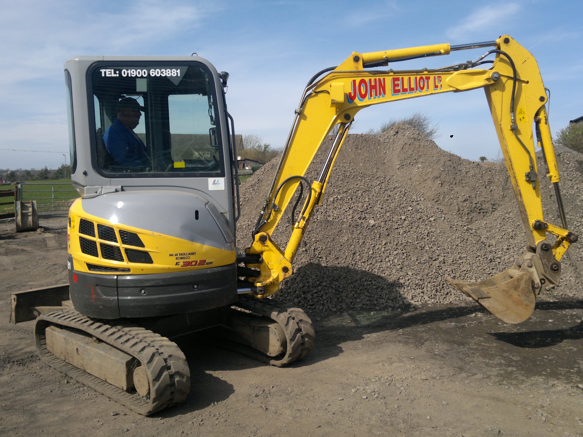 Yellow mini excavator with John Elliot Ltd on the boom loading gravel