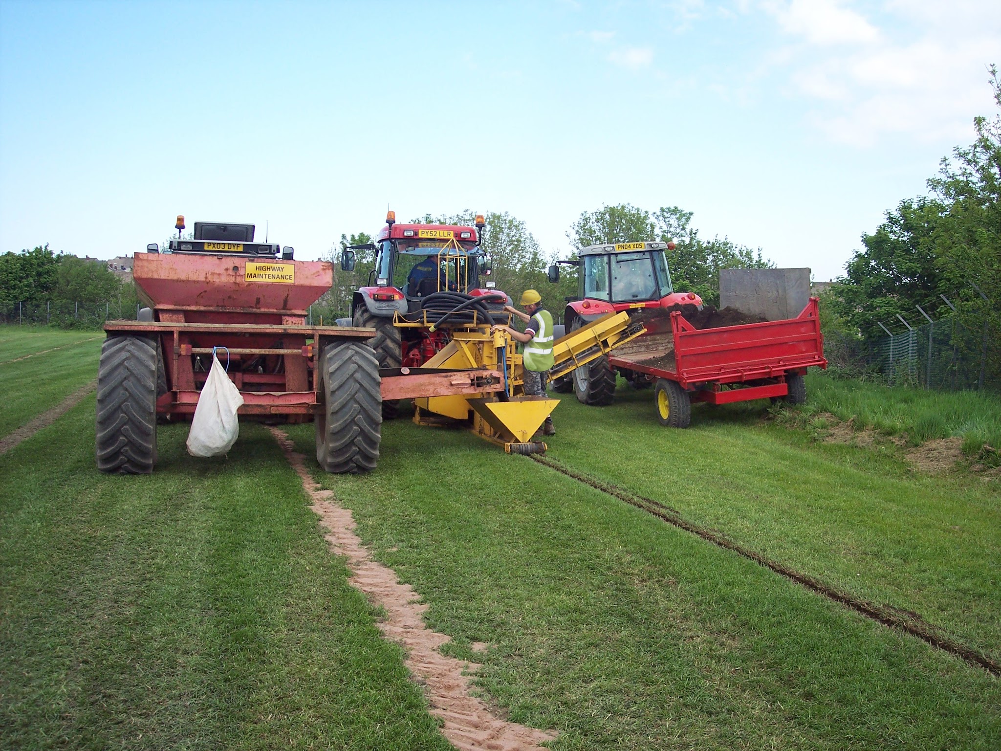 Tractor-drawn trencher and gravel trailers laying drains across a grass pitch